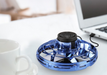 A blue, circular Mini Drone LED UFO Type Flying Helicopter Spinner Fingertip by Maroon Asteria with RGB lights and a protective grid cover is placed on a white table next to a white coffee cup and an open laptop. The drone is connected to the laptop via a black USB cable, creating a cozy work environment. The background is slightly blurred.