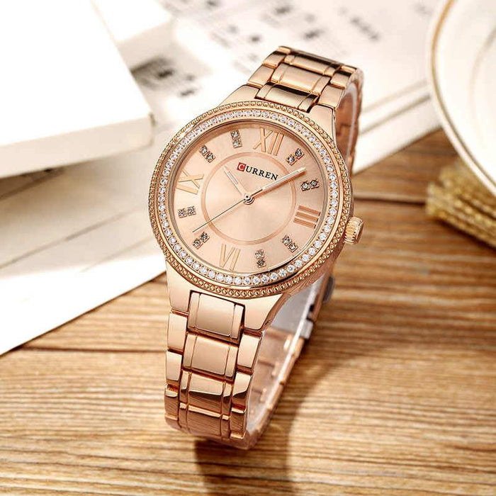 The CASSANDRA Women's Classic Watch by Lilac Quartz, with its elegant rose gold finish and round face, showcases Roman numerals and gemstone hour markers. Situated on a wooden surface amidst open books and a white plate, this watch is powered by precise Japan Quartz movement, embodying both sophistication and accuracy.