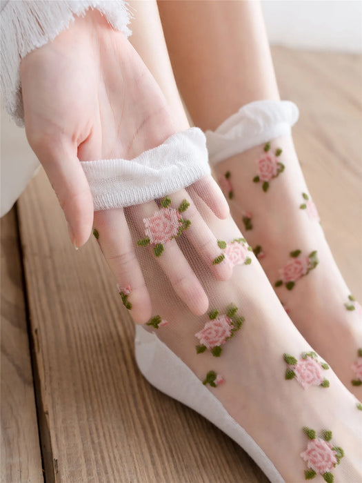 A hand adjusts a sheer sock, embroidered with pink roses and green leaves. The Lacatang Shop's Vintage Floral Lace Ruffle Socks, designed in a Kawaii Harajuku style, embellish the foot against wooden flooring, with a white garment sleeve partly visible.