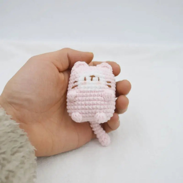 A person is holding a Cute Cat Crochet Keychain from Lacatang Shop in their hand. The small, pink, crocheted plush toy shaped like a cat has a white face with simple stitched details for the eyes, nose, and mouth. The adorable knitted keyring makes for a perfect accessory for car keys or as a bag pendant. The background is white and the person's sleeve is partially visible.