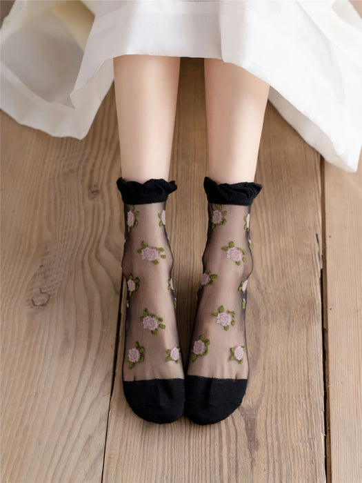 A person wears Lacatang Shop's Vintage Floral Lace Ruffle Socks in sheer black with pink embroidery, seated on a wooden floor. The scene evokes a Kawaii Harajuku vibe, with a glimpse of a white skirt at the top of the image.