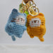 Two Cute Cat Crochet Keychains from Lacatang Shop, one in blue and one in yellow, each with adorable embroidered facial features and soft whiskers, are hanging from a gold keyring against a plain background.