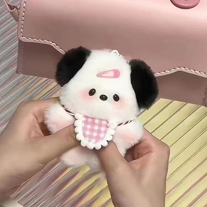 A person holds the Cute Plush Puppy Keychain from Lacatang Shop, adorned with pink cheeks, a tiny pink party hat, a checkered bib, and black ears. Ideal as a birthday gift for women and girls, it's accompanied by a light pink purse charm on a striped surface.