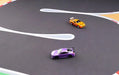 Two RC mini cars, one purple and one orange, race on a Lacatang Shop Portable 1/76 Scale Turbo Racing Rubber Mat, complete with painted lines and red-and-white striped curbs for a realistic track experience.