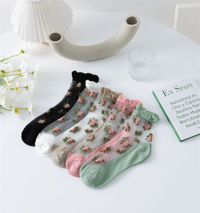 A set of six pairs of Lacatang Shop's Delicate Vintage Floral Lace Ruffle Socks, featuring transparent ankle designs in black, white, beige, pink, and green, is displayed on a white table with nearby white flowers, a decorative piece, and an open book.