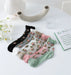 A set of six pairs of Lacatang Shop's Delicate Vintage Floral Lace Ruffle Socks, featuring transparent ankle designs in black, white, beige, pink, and green, is displayed on a white table with nearby white flowers, a decorative piece, and an open book.