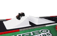 A Lacatang Shop toy car speeds on a white ramp, part of the customizable TURBO RACING 1:76 Remote Control Car Track Mat with cement blocks and drift jumps. The mat has red and gray borders with "TURBO RACING" on a green section.