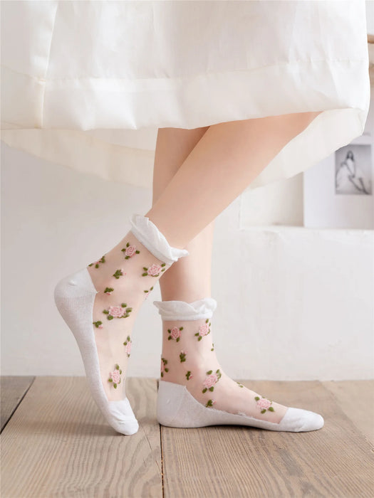 Someone wearing Lacatang Shop's Vintage Floral Lace Ruffle Socks, featuring embroidered pink flowers and green leaves, stands on a wooden floor. Paired with a white knee-length skirt, the scene exudes a light and airy feel reminiscent of transparent crystal silk elegance.