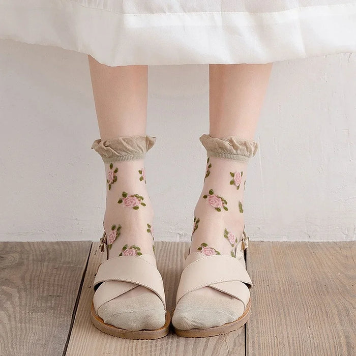 A person stands on a wooden floor, wearing a white skirt and Lacatang Shop's Vintage Floral Lace Ruffle Socks adorned with pink roses in kawaii Harajuku style, paired with beige sandals.