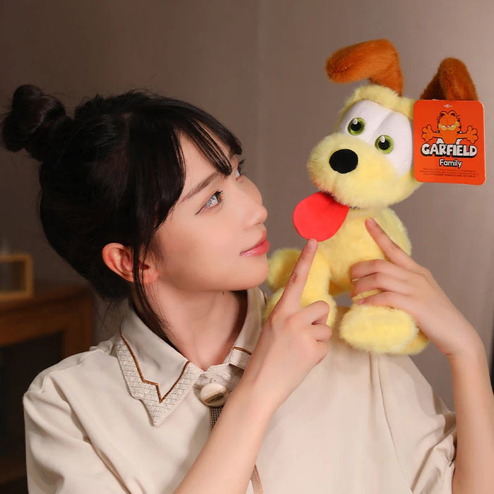 A person in a beige shirt is holding and playfully pointing at a Lacatang Shop Kawaii Oudi dog plush toy with floppy brown ears, large eyes, and a red tongue sticking out. This cute toy from "Kawaii Oudi Dog & Ugly Cat Plush Toys" collection is perfect for any plush toys enthusiast.