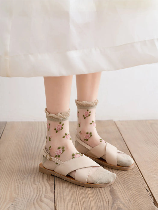 A person is wearing Lacatang Shop's Delicate Vintage Floral Lace Ruffle Socks with Crystal Silk, styled in kawaii Harajuku fashion, paired with beige criss-cross sandals and a white flowing skirt while standing gracefully on a wooden floor.