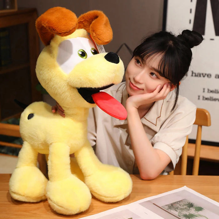 A woman with a bun is seated indoors, smiling at a large Kawaii Oudi Dog plush toy from Lacatang Shop. The adorable stuffed dog has floppy ears, big eyes, and a red tongue, accompanied by books and framed pictures in the background. Ideal for room décor and perfect as gifts for kids.