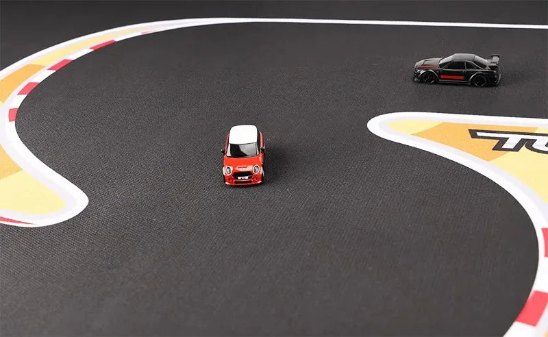 Two 1:76 RC mini cars race on a Lacatang Shop's Large 160x90cm Portable Turbo Racing Track Mat. A red and white car takes a curve in the foreground with a black car following. The track features a dark, high-grip surface with white and red edging, complemented by yellow markings.