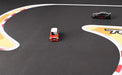 Two toy cars, one red and white and the other black with red accents, race on Lacatang Shop's Large Portable Turbo Racing Rubber Mat. The track features a sharp curve with red and white stripes against a dark textured background, ideal for 1:76 RC Mini Cars.