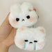 A hand is holding two fluffy white Cartoon Plush Rabbit Doll Toy Keychains, crafted from high-quality plush material and shaped like animal faces. The top plush, featuring an indifferent expression with small eyes and a slight frown, and the bottom plush, depicting a sad expression with tiny eyes and blue tear streaks down its face, are part of Lacatang Shop's Sweet Cute Bag Pendant Charms collection for women and couples.