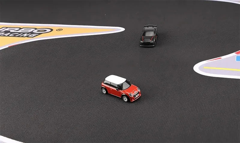 Two small toy cars are on a Lacatang Shop Large Portable Turbo Racing Rubber Mat, featuring colorful markings. A red car with a white roof leads while a black car follows, mimicking a miniature racing setup for 1:76 RC mini cars.