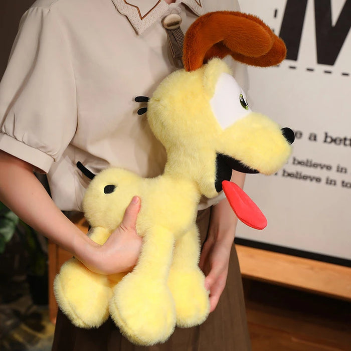 A person holds a large yellow plush toy resembling the Kawaii Oudi Dog from Lacatang Shop, featuring big eyes, brown ears, and a floppy red tongue. They wear a light shirt and brown pants against a backdrop with a framed picture or poster.