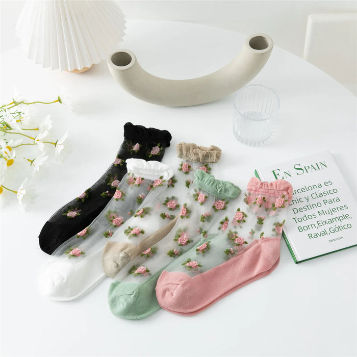 Lacatang Shop's Delicate Vintage Floral Lace Ruffle Socks with Crystal Silk for Women and Girls are displayed on a table in black, white, beige, and pink. A white vase, a glass of water, and the green book "EN SPAIN" enhance the charming setup adorned with white flowers.