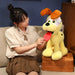 A person in a beige shirt and brown skirt sits on a sofa, admiring a large Kawaii Oudi Dog plush toy from Lacatang Shop. The yellow stuffed doll with brown ears and a red tongue is placed on a wicker basket against the dark wall, enhancing the cozy atmosphere.