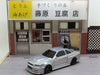 A Lacatang Shop 2.4G RC Drift Car 1/43, a high-speed, four-wheel-drive model with a silver body and black rear spoiler, is parked in front of a meticulously detailed miniature storefront. The storefront showcases Japanese signage and appears to be selling tofu and fried tofu. Brick and wooden wall details in the background add an extra layer of charm to this captivating scene.