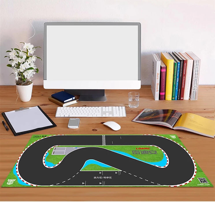 A wooden desk holds a computer, keyboard, mouse, small plant, books, and an open magazine. In the foreground is a Lacatang Shop's LDARC V64 miniature 1/64 scale RC car racing track mat (2400mm x 1200mm) with a winding design.