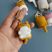 A hand holds a Cute Cat Crochet Keychain from Lacatang Shop, featuring a small animal with a yellow body and white face. Other similar knitted keychains in various colors are scattered in the background. The keychain is attached to a zinc alloy ring.