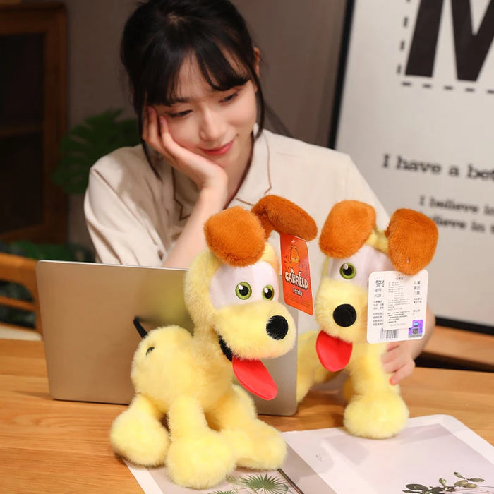 A person smiles at two Lacatang Shop kawaii Oudi Dog plush toys with big eyes and brown ears, long red tongues, and tags while sitting at a desk with an open laptop; a framed picture is partly visible on the wall.