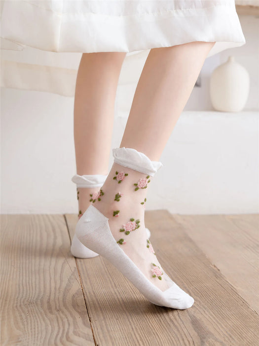 A person wears Lacatang Shop's Vintage Floral Lace Ruffle Socks for Women, featuring pink embroidery and white frilled cuffs on a wooden floor, with a white dress partially visible. The cream-colored room's subtle decor enhances the kawaii Harajuku style ambiance.