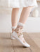 A person wears Lacatang Shop's Vintage Floral Lace Ruffle Socks for Women, featuring pink embroidery and white frilled cuffs on a wooden floor, with a white dress partially visible. The cream-colored room's subtle decor enhances the kawaii Harajuku style ambiance.