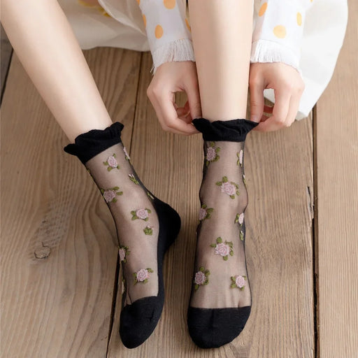 A person dressed in a white dress with orange polka dots sits on a wooden floor, wearing Lacatang Shop's Delicate Vintage Floral Lace Ruffle Socks. These kawaii Harajuku style crew socks feature embroidered pink flowers and black toes and heels.