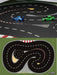 Two remote control toy cars race on a winding track with red and white borders. The TURBO RACING 1:76 Track Mat by Lacatang Shop features sharp turns, marked lanes, cement blocks, and drift jumps, with the "Turbo Racing" logo visible in the corner.