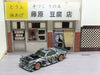 A 2.4G RC Drift Car 1/43 4WD Remote Control Car from Lacatang Shop, featuring a zebra-striped design and orange wheels, is placed on the ground in front of a miniature building adorned with Japanese signage. The building includes a sliding glass door and a window with bars, along with a yellow awning and a wooden fence.