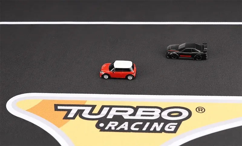 Two 1:76 RC Mini Cars race on the black Lacatang Shop Racing Mat. A red car with a white roof leads a black car with red accents. The track displays a yellow and white "Turbo Racing" logo on the Large Portable Turbo Racing Rubber Mat from Lacatang Shop.