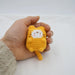 A hand holding a Cute Cat Crochet Keychain from Lacatang Shop, showcasing a small, yellow cat plushie with a white face and brown stripes. The round cat figure features tiny ears, a long tail, and short paws, resembling an adorable knitted keychain. It is set against a plain white background.