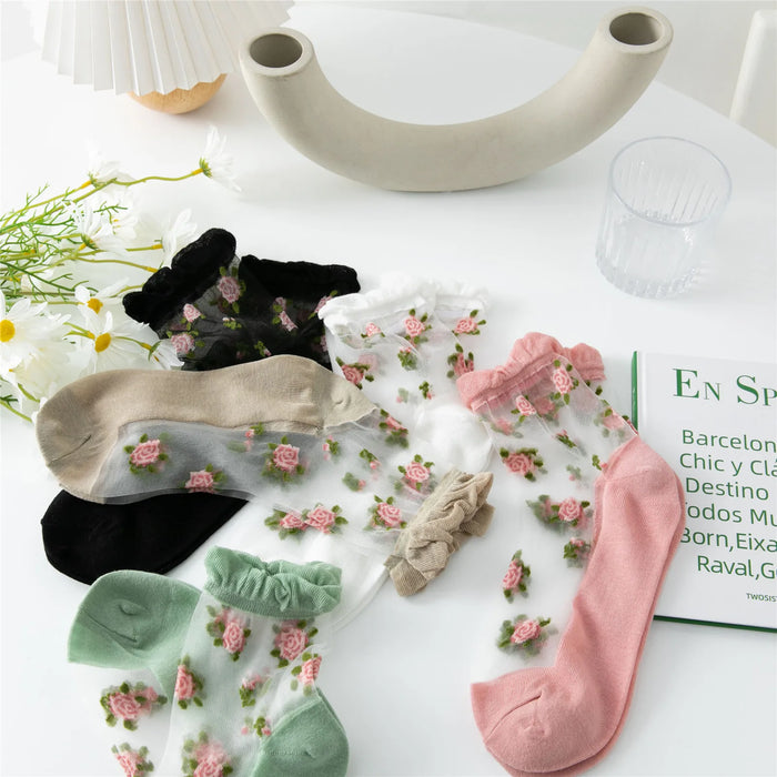 On a white surface, Delicate Vintage Floral Lace Ruffle Socks with Crystal Silk for Women and Girls by Lacatang Shop are displayed, embodying the Kawaii Harajuku style with pastel cuffs. Nearby are daisies in a vase, a clear glass, an open book, and a modern curved decorative piece.