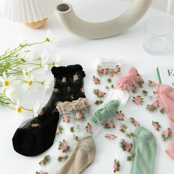 The Delicate Vintage Floral Lace Ruffle Socks from Lacatang Shop are Kawaii Harajuku Style crew socks for women and girls, featuring colorful sheer designs in black, white, green, and pink. They're displayed on a white surface with daisies, a curved decor piece, a glass of water, and a book.