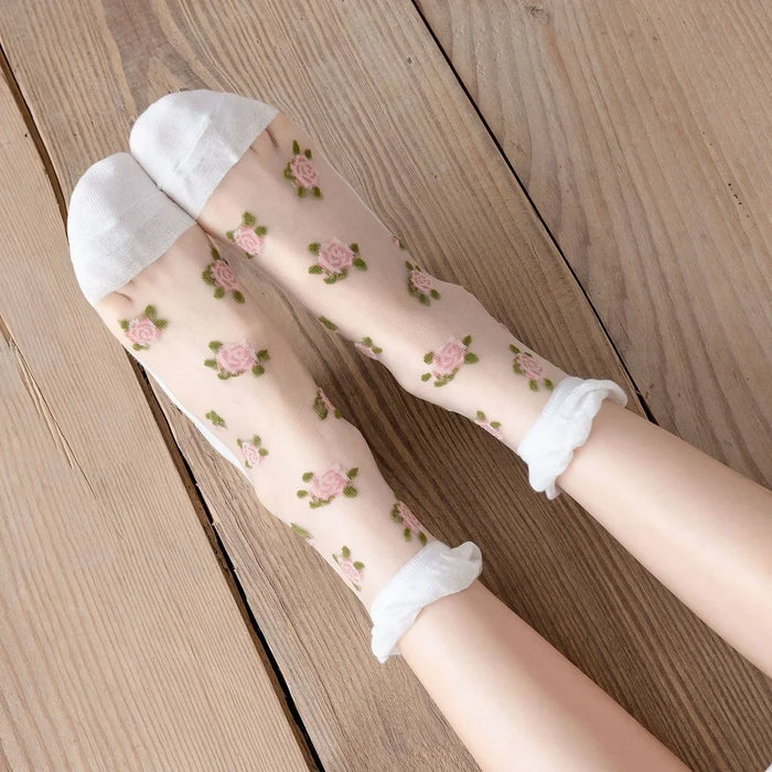 Feet adorned in Lacatang Shop's Vintage Floral Lace Ruffle Socks, featuring sheer pink roses and green leaves, rest on a wooden floor. These Kawaii Harajuku style socks charm with white toes and cuffs.