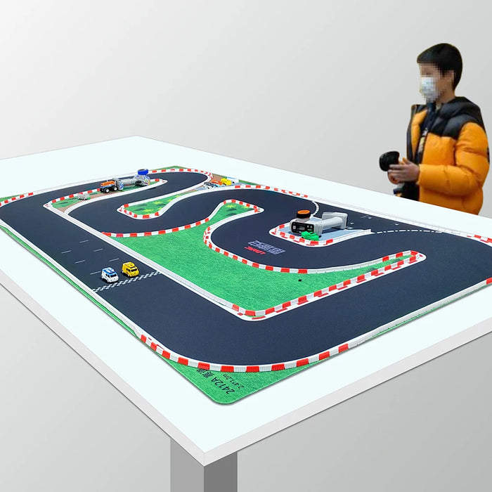 A person in a yellow jacket operates a remote beside the Lacatang Shop Portable PK Mesh Race Track Mat, featuring red and white barriers. Mini RC cars are lined up along the winding black track on a green surface, creating an immersive racing experience.