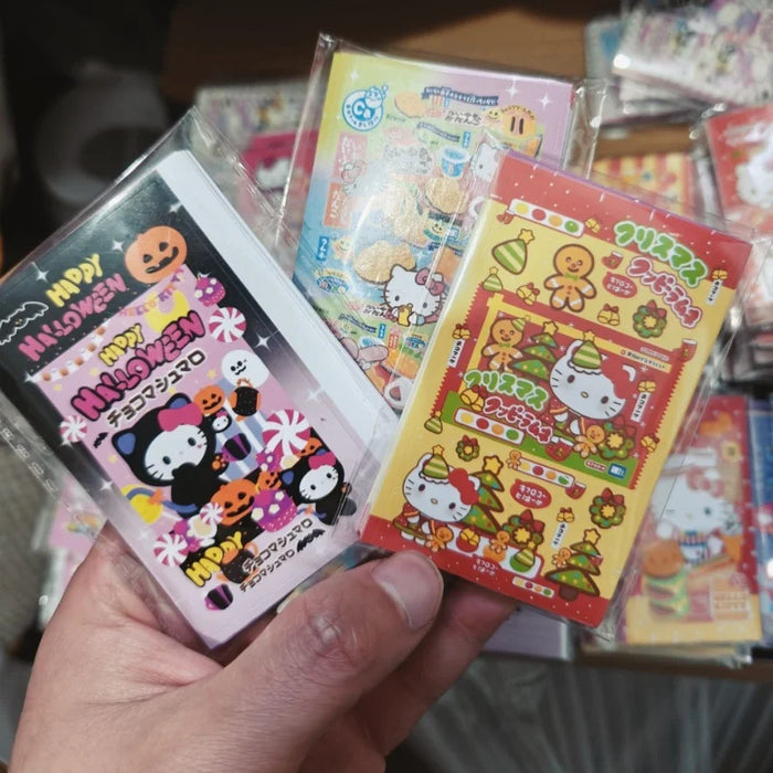 A person holds Japanese candy packs adorned with colorful Halloween designs featuring cartoons and pumpkins. Sanrio Characters add a delightful touch amidst the whimsy. In the background, more candy packs lie alongside Lacatang Shop’s Kawaii Hello Kitty Sealing Stickers for ages 8+.