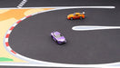 Two small toy cars, purple and orange, race on Lacatang Shop's Portable Turbo Racing Rubber Mat for 1:76 RC Mini Car Track. The purple car leads near the checkered section with the orange in pursuit. This mat features distinct white lines and vibrant sections for thrilling mini races.