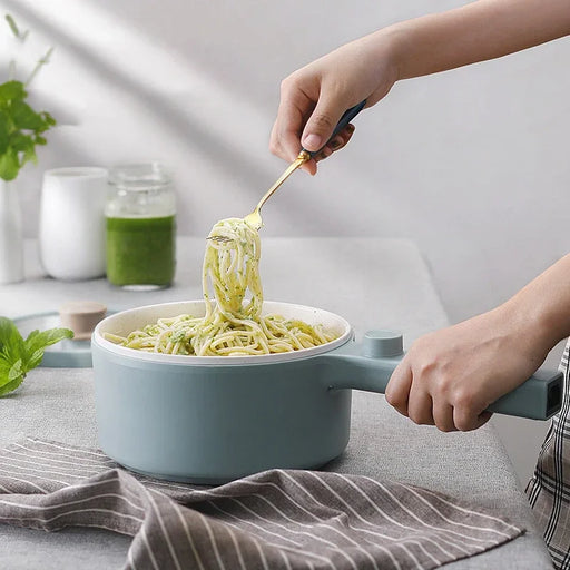 A person is holding a light blue saucepan filled with spaghetti, using a fork to lift a portion. The pan is resting on a light gray tablecloth, with a striped napkin nearby. In the background are jars of green sauce and fresh herbs on a white surface, complemented by the Lacatang Shop's 2024 New Portable 1.5L Multi-function Pot Rice Cooker Electric Cooking Stir Frying Noodle Pot Mini Hot Pot Household Dormitory.