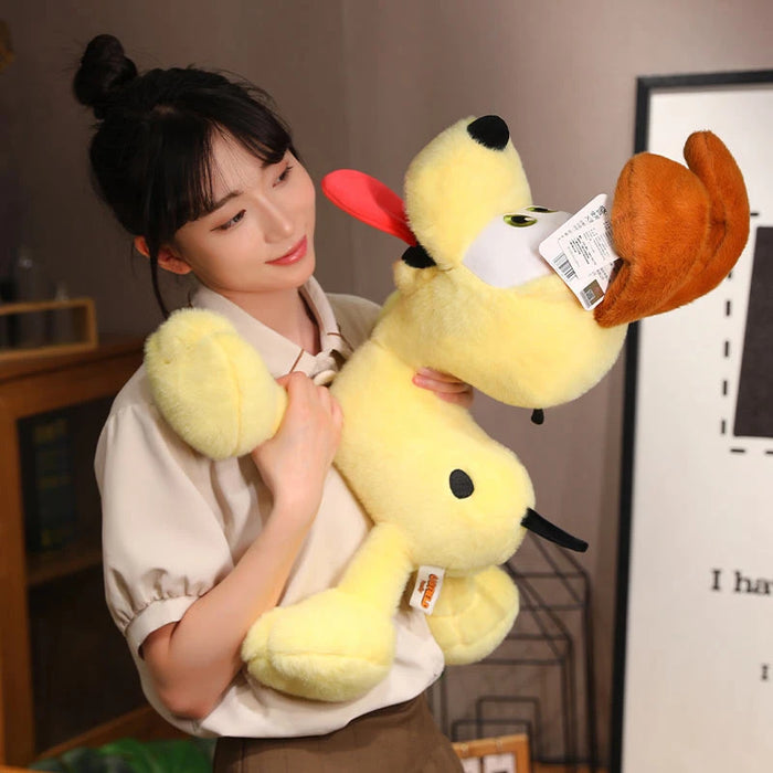 A dark-haired person smiles while holding a large, yellow Kawaii Oudi Dog Plushie from Lacatang Shop, featuring floppy ears and a red tongue. The setting is indoors with a shelf and framed picture, exuding a cute and quirky design vibe.