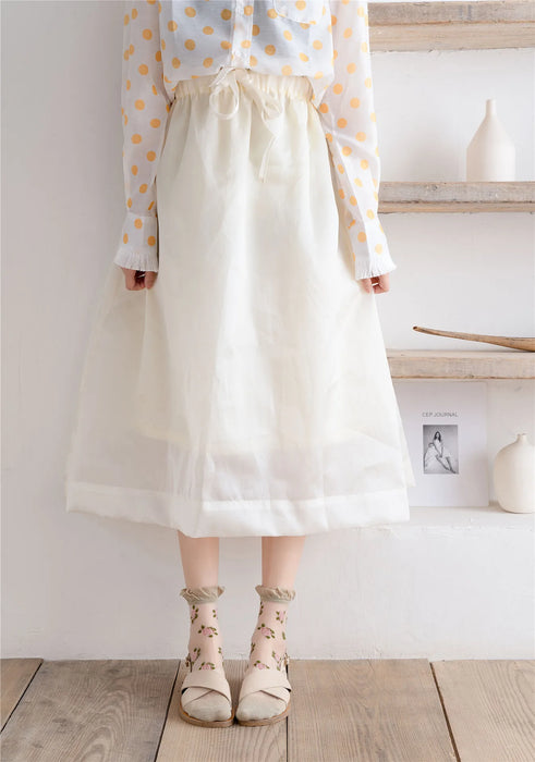 A person stands on wooden steps, wearing a sheer white skirt, polka dot blouse, and Lacatang Shop's Delicate Vintage Floral Lace Ruffle Socks with Crystal Silk in kawaii Harajuku style. The minimalist decor features vases and a small photo on stacked shelves.