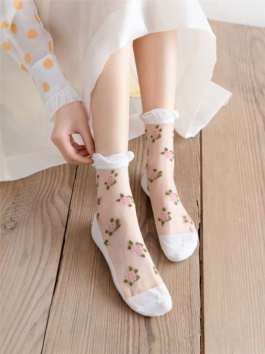 A person in a white dress sits on a wooden floor, adjusting their transparent Kawaii Harajuku style socks from Lacatang Shop. The Vintage Floral Lace Ruffle Socks feature pink floral embroidery and white cuffs. Their dress has sheer sleeves with orange polka dots, adding whimsy to the scene.