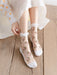 A person sits on a wooden floor wearing Lacatang Shop's Delicate Vintage Floral Lace Ruffle Socks with Crystal Silk, adorned with pink roses and white toe caps. They adjust one sock while dressed in a flowing skirt and a polka-dotted white blouse, embracing kawaii Harajuku style.