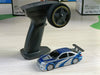 A sleek black remote control lies on a table next to the 2.4G RC Drift Car from Lacatang Shop, a small blue and silver high-speed racing car equipped with a rear spoiler. In the background, there are indistinct boxes. The remote features a prominent wheel and several buttons, perfect for controlling the high-speed thrills of your new 1/43 4WD Remote Control Car.