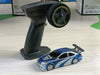 A detailed miniature blue 2.4G RC Drift Car from Lacatang Shop, featuring silver stripes and a black spoiler, is displayed on a light wooden surface. To the left of the car sits its black remote control with a large circular steering wheel and several buttons, used to operate this high-speed, four-wheel drive radio-controlled racing car model.