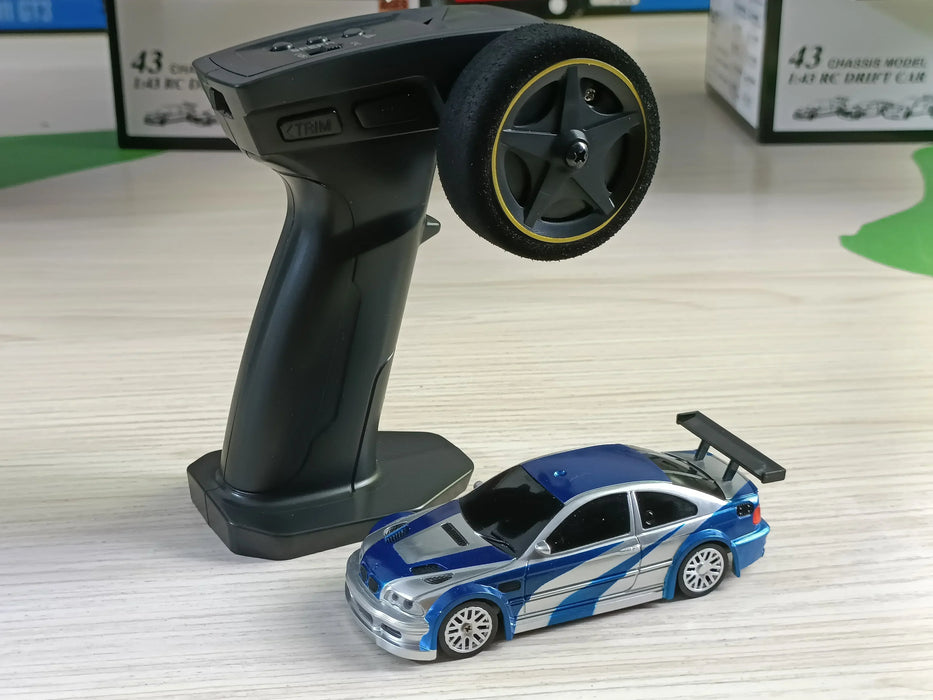 A black remote control is positioned upright next to a small blue and silver 2.4G RC Drift Car 1/43 4WD from Lacatang Shop on a light-colored wooden surface. The high-speed car features a spoiler and racing decals. The background includes boxes partially visible in a blurred view.