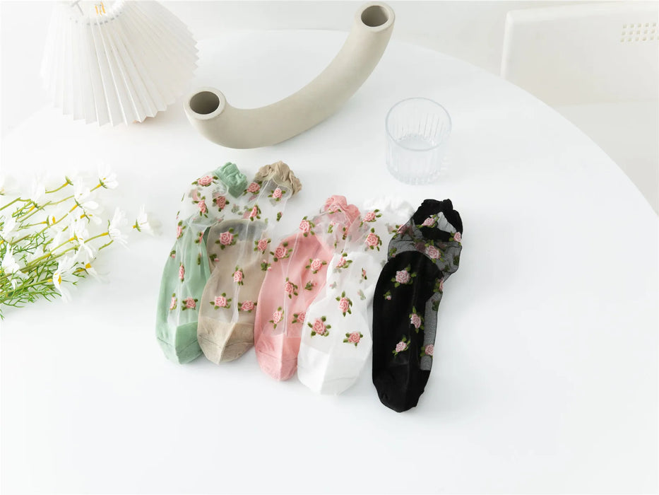 A set of Vintage Floral Lace Ruffle Socks from Lacatang Shop, available in green, beige, pink, white, and black, are showcased on a round white table beside a decorative curved item, a crystal-clear glass of water, a pleated lampshade, and white flowers.