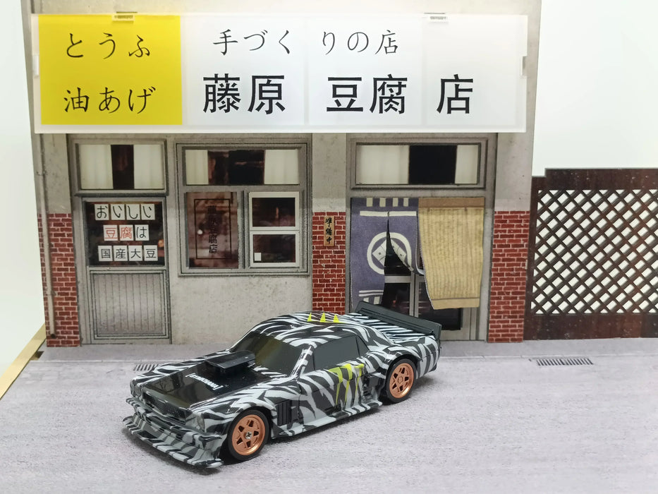 The 2.4G RC Drift Car 1/43 by Lacatang Shop, featuring a black and white camouflage pattern and bronze wheels, is placed in front of a model Japanese tofu shop. The shop showcases a yellow and white sign with Japanese text above the entrance and wooden panels on one side.
